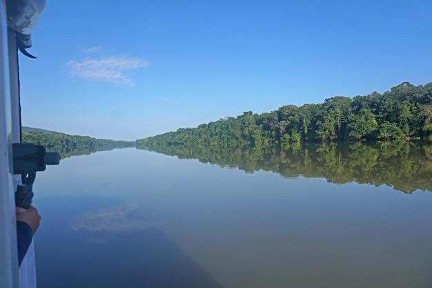 A panoramic view of the Amazon River with a glimpse of the rainforest in Brazil, showcasing the lush greenery, various boats navigating the river, and clear skies above, encapsulating the vastness and natural beauty of the Amazon basin.