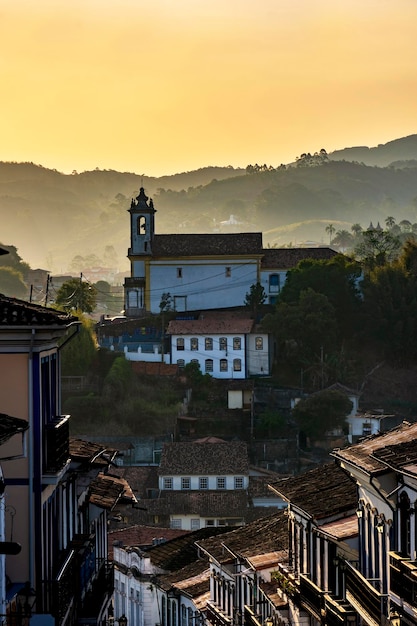 A picturesque view of Ouro Preto, a historical city in Minas Gerais, Brazil, showcasing its colonial architecture, cobblestone streets, baroque churches, and rolling green hills in the background under a clear blue sky.