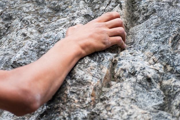 A close-up shot captures a climber's hands gripping a rocky cliff face. The climber is wearing climbing gloves, and the rock is rough and textured. The background is blurred, highlighting the climber's focus and determination.