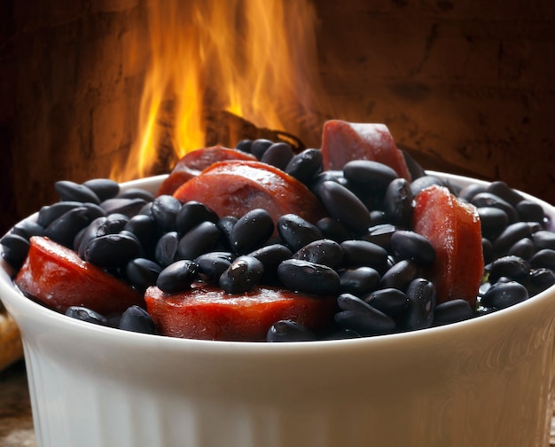 A close-up shot of a steaming bowl of feijoada, showcasing the rich, dark color of the beans and the various cuts of meat. Garnishes of orange slices, collard greens, and farofa are arranged around the bowl.