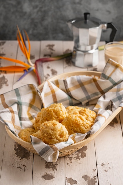 A pile of freshly baked pão de queijo, golden brown and slightly puffed, spilling out of a rustic basket. The background features a traditional Brazilian kitchen setting.