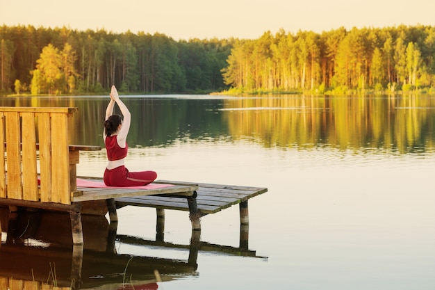 An outdoor yoga session taking place on a wooden platform overlooking a serene lake at sunrise. Participants are engaged in various yoga poses, guided by an instructor.