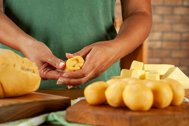 A close-up shot of a woman's hands carefully preparing a batch of pão de queijo dough on a floured wooden surface. The ingredients, such as cheese, tapioca flour, eggs, and milk, are neatly arranged around the dough. The focus is on the texture and details of the dough being kneaded, conveying a sense of tradition and care.
