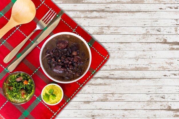 An overhead shot showcasing a table setting with a complete feijoada meal. It includes a bowl of feijoada, a plate of white rice, sautéed collard greens, farofa, orange slices, and a small bowl of hot sauce. The presentation is appealing and emphasizes the variety of components that make up the dish.