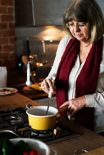 Medium shot of an older woman stirring a large pot of simmering food, likely a traditional stew, in a rustic kitchen setting, sunlight streaming in through a window.