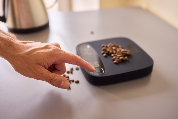 A close-up shot of a person using a small, precise kitchen scale to weigh chocolate for a brigadeiro recipe. The scale is on a clean, stainless steel countertop, with other ingredients like condensed milk and cocoa powder visible in the background. Soft, focused lighting highlights the numbers on the scale and the texture of the chocolate.