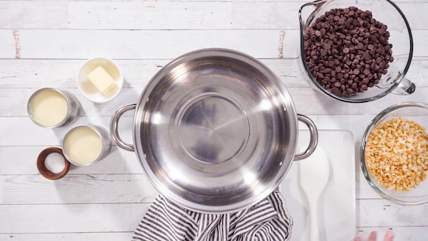 A top-down view of a saucepan on a stove, filled with brigadeiro mixture being stirred with a wooden spoon. The mixture is smooth and glossy, with visible steam rising. Ingredients like chocolate bars, condensed milk cans, and butter are arranged around the stove. Warm, inviting lighting.