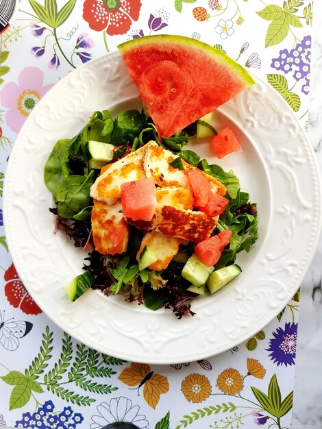 A bright, overhead shot of a summer salad made with watermelon, grilled halloumi cheese, and fresh mint. The salad is arranged on a white plate, and the vibrant colors of the ingredients are highlighted by natural sunlight.