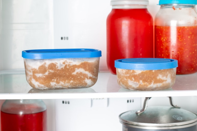 Close-up of labeled containers filled with various leftovers (cooked chicken, roasted vegetables, rice), neatly organized inside a refrigerator.