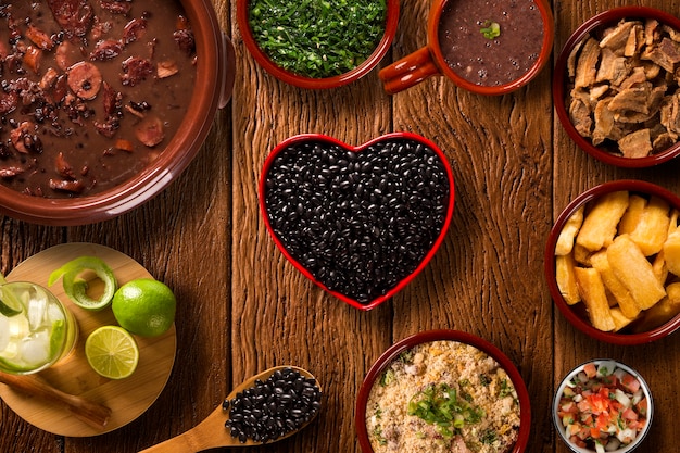 Close-up of various ingredients for feijoada laid out on a table: black beans, different cuts of salted pork, sausages, and spices, each in its own small bowl. The focus is on the textures and colors of the raw ingredients, with natural light enhancing the details.